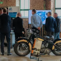 Grupa uczestników rozmawia przy stoisku ITA z&nbsp;monitorem i&nbsp;zabytkowym motocyklem.