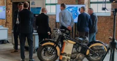 Grupa uczestników rozmawia przy stoisku ITA z&nbsp;monitorem i&nbsp;zabytkowym motocyklem.